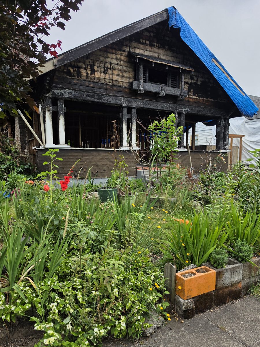 Front of the house after the fire, with charred porch, siding and gable under a blue tarp